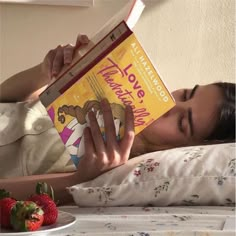 a woman laying in bed reading a book with strawberries on the table next to her