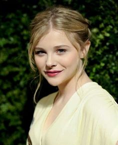 a woman with blonde hair wearing a white shirt and smiling at the camera while standing in front of some bushes