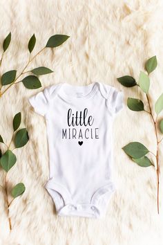 a white bodysuit with the words little miraacie on it next to some green leaves