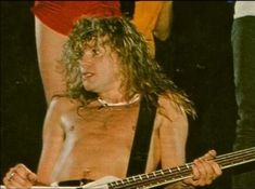 a shirtless man with long hair holding a guitar in front of other men and women