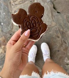 someone holding up a mickey mouse cookie in their left hand and another person's feet on the ground behind them