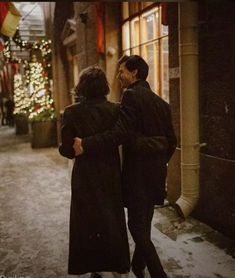 two people walking down an alley way in the snow with christmas decorations on the wall behind them