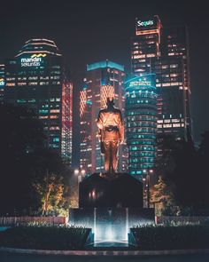 a large statue in front of some tall buildings at night with the city lights behind it