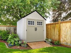 a small shed in the middle of a yard with flowers and plants around it, next to a wooden fence