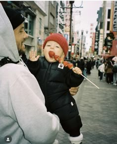 a man holding a baby in the middle of a crowded street with food on sticks