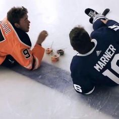 two young men sitting on the ice and one is holding his hand out to him