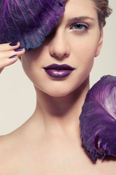 a woman with purple lipstick holding a flower in front of her face and looking at the camera