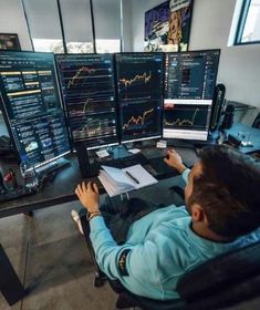 a man is sitting in front of three computer screens with graphs on them and working on his laptop