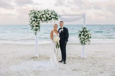 a bride and groom standing on the beach under an arch decorated with white flowers for their wedding ceremony