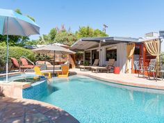 an outdoor pool with chairs and umbrellas next to the side of a house that is surrounded by trees