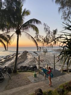 a woman holding a surfboard next to the ocean at sunset with palm trees in the foreground