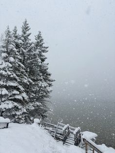 snow covered trees and benches on the side of a snowy hill with water in the background