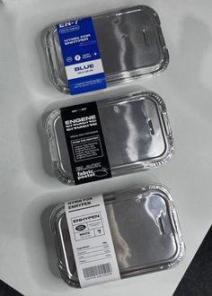 two empty trays sitting next to each other on a white counter top, one has a blue label and the other is silver