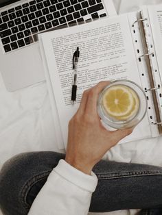 a person holding a glass with lemon in front of an open book and laptop computer