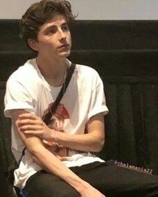 a young man sitting on top of a black couch next to a white wall and wearing a white t - shirt