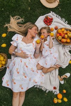 two women laying on the grass with apples and oranges