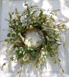 a wreath with white flowers and green leaves hanging on the side of a house door