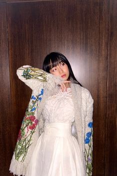 a woman standing in front of a wooden wall wearing a white dress and jacket with flowers on it