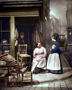 two women are sitting on the sidewalk in front of an old fashion shop, talking to each other