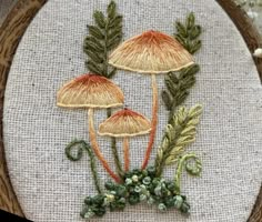 two mushrooms with leaves and acorns are on a linen background in an embroidery hoop