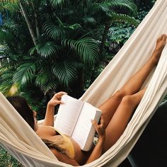 a woman laying in a hammock reading a book with her legs crossed and holding an open book