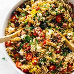 a bowl filled with pasta salad topped with tomatoes and feta cheese, next to a wooden spoon