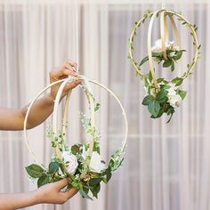 two circular hanging flower baskets with greenery and white flowers are held up by someone's hands