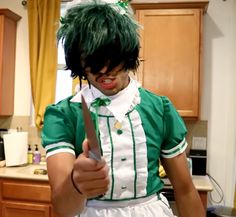 a man dressed in green and white holding a knife with his right hand while standing in a kitchen