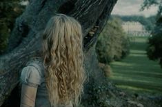 a woman with long blonde hair standing in front of a tree and looking at the ground