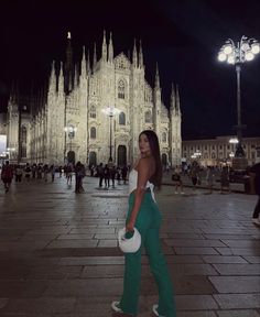 a woman is walking in front of a cathedral at night with her hand on her hip