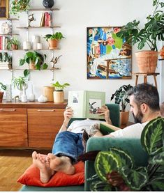 a man sitting on a couch reading a book in front of a plant filled wall