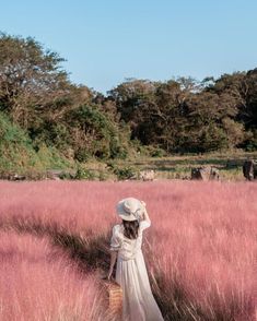 a woman in a white dress and hat walking through tall grass with an elephant behind her