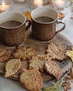 two mugs of coffee and some cookies on a wooden board with leaves around them