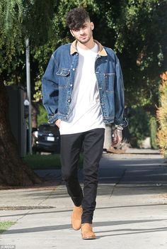 a young man walking down the street wearing a jean jacket and black pants with brown suede boots