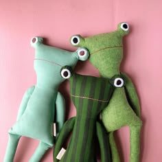 three green stuffed animals sitting next to each other on a pink surface with eyes and legs