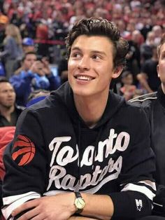 a young man sitting in front of a crowd at a basketball game wearing a toronto raptors hoodie