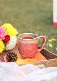 a cup of tea on a tray with flowers and pine cones next to it in the grass