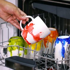 a person is pouring something into a dishwasher full of colorful mugs and cups