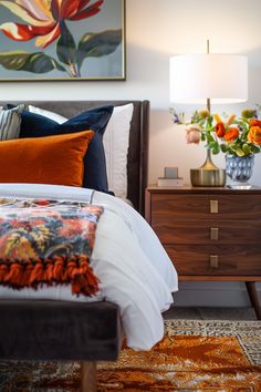 a bed with orange and blue pillows on top of it next to a nightstand filled with flowers