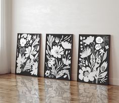 three black and white floral paintings sitting on top of a hard wood floor next to a window