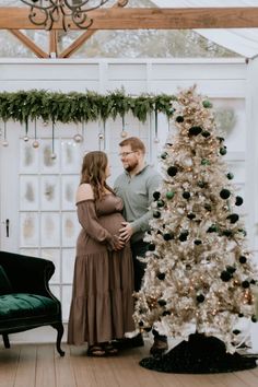 a pregnant couple standing next to a christmas tree