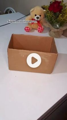 a teddy bear sitting in a cardboard box on a table next to a vase with flowers