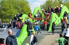 many people are walking around in the park and on the playground slide, while one person is riding a skateboard