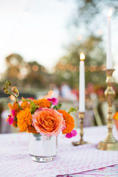 an orange and pink flower arrangement in a silver vase on a white table cloth with candles