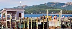 some people are standing on the dock by the water and buildings with mountains in the background