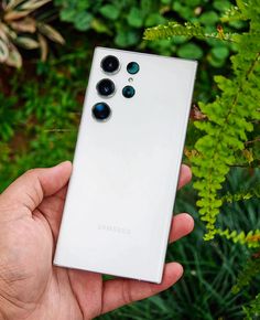 a hand holding a white samsung phone in front of green plants and bushes, with the back camera facing up