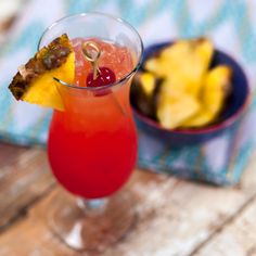 a red drink with pineapple and cherries on the rim next to a bowl of fruit