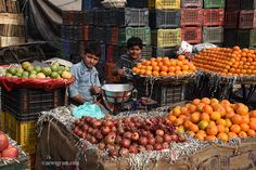 two men standing in front of baskets of oranges and other fruit on display at an outdoor market