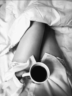 a woman laying in bed with her legs crossed and holding a cup of black coffee