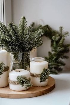 some white candles are sitting on a wooden tray next to evergreen branches and pine cones